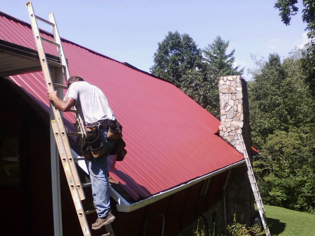 Roofer on a ladder beside a red metal roof and stone chimney — Soffit & Fascia Repair by Roofing Brothers in Naval Air Station Jrb